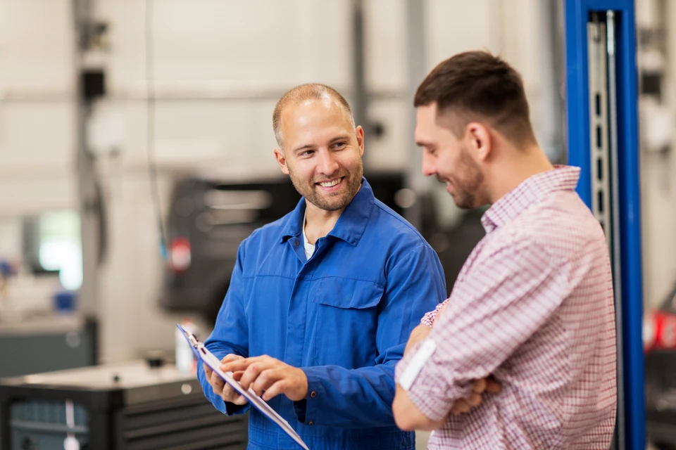 Atelier automobile avec mécanicien en combinaison bleue souriant tenant une tablette numérique, discutant avec un client en chemise à carreaux